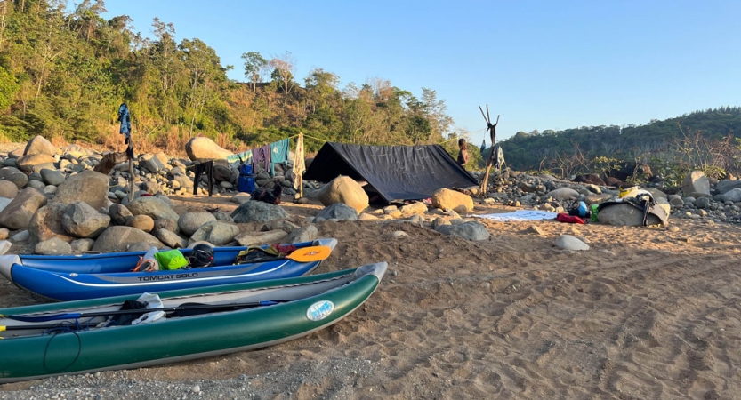 A tarp shelter and watercraft rest on the shore of a river near trees